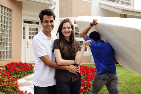 new homeowners standing happily in front of home while mover brings in furniture