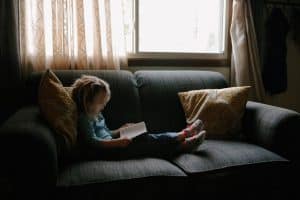 Little girl sitting on grey sofa reading a book
